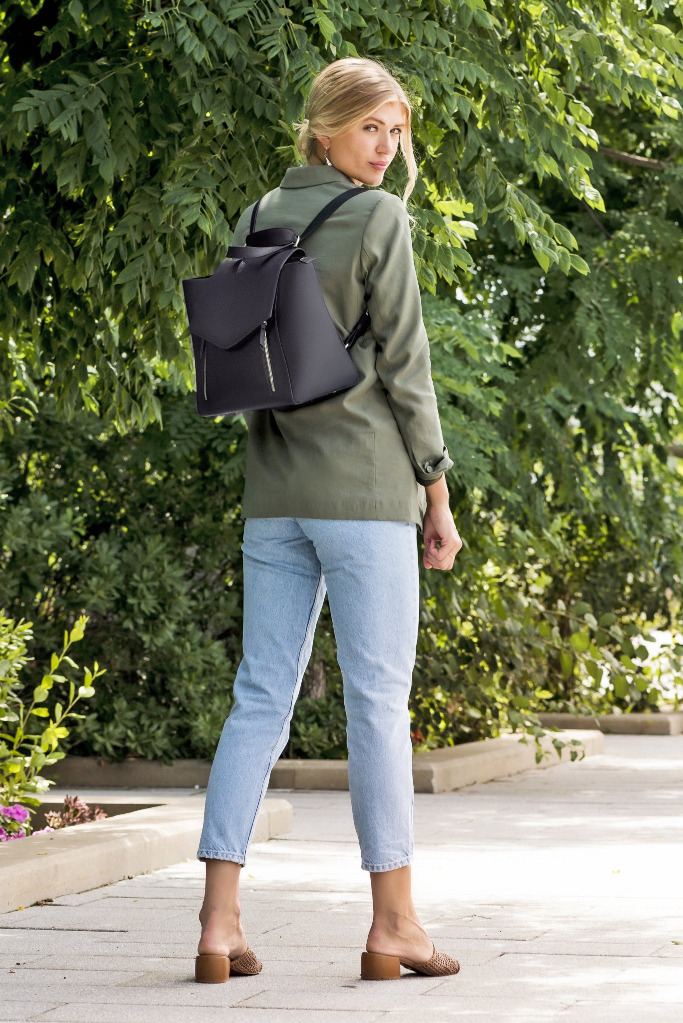 Woman wearing AGIKI Classic Black convertible leather laptop work bag for women as a backpack, outdoors with greenery.